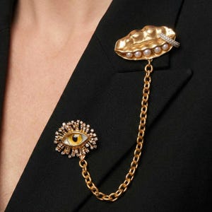 Può includere: Set di spille dorate. Una spilla a forma di occhio con iride gialla e una a forma di labbro con perle, collegate da una catena dorata. Entrambe le spille sono decorate con piccoli cristalli. Il set è esposto su un blazer nero.