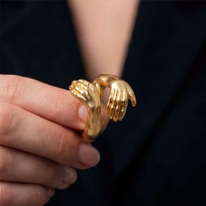 Könnte beinhalten: Ein goldfarbener Ring mit zwei ineinander verschlungenen Händen. Der Ring ist ein einzigartiges Schmuckstück mit detaillierten Handskulpturen, ideal, um jedem Outfit einen Hauch von künstlerischem Flair zu verleihen. Der Ring besteht aus glänzendem Metall.