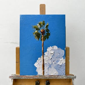 May include: An oil painting on canvas featuring a single palm tree centered against a vibrant blue sky. The tree has a textured brown trunk and green fronds. A large, thick-impasto white cloud sits at the base of the tree, adding depth.
