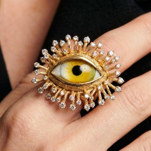 May include: A gold-toned ring featuring an eye design with a yellow iris and black pupil. The eye is surrounded by a sunburst of small, clear gemstones. The ring is set on a finger against a dark background.