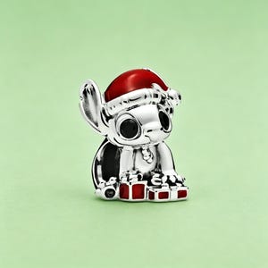 May include: A silver-colored charm featuring a cartoon character wearing a red Santa hat. The character has large black eyes and is holding several small red gift boxes. The charm is set against a light green background.