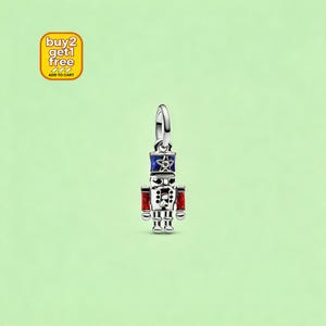 May include: A silver charm shaped like a nutcracker figure, featuring a blue hat with a star detail, red glittery accents on the arms, and black enamel detailing on the face and body. The charm includes a circular bail at the top for attachment.