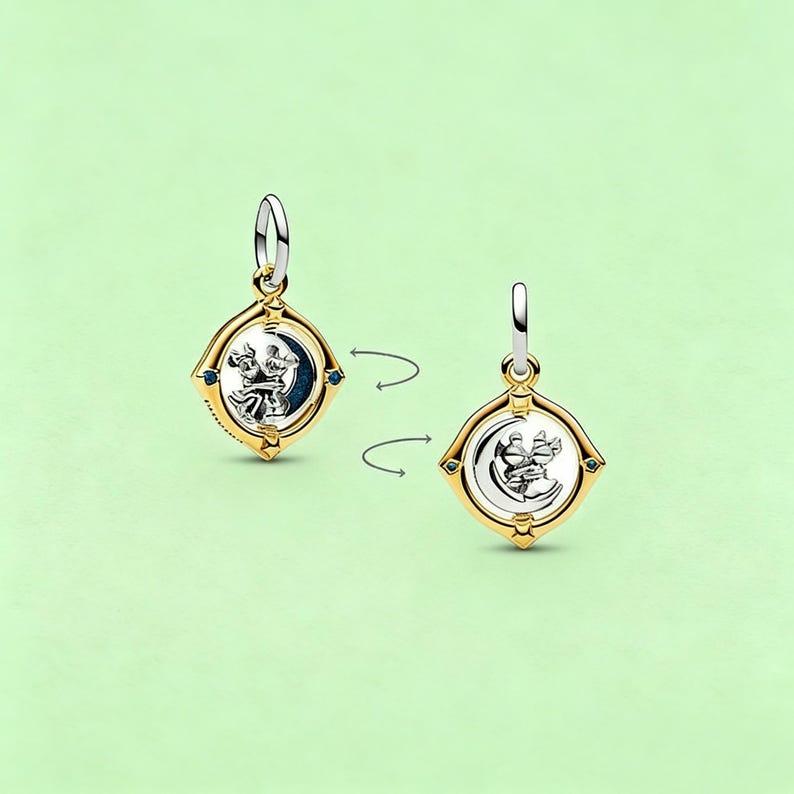 May include: Two-sided gold-toned pendant with a silver-colored design of a couple dancing in front of a crescent moon. The pendant has a square shape with blue accents and a silver-colored bail.