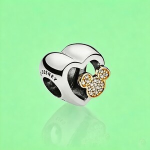 May include: A silver heart-shaped charm bead featuring the word DISNEY engraved on the side. A gold-toned Mickey Mouse head silhouette encrusted with small clear stones is attached to the front of the heart, creating a layered metallic design.
