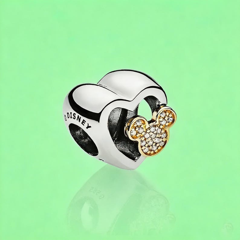 May include: A silver heart-shaped charm bead featuring the word DISNEY engraved on the side. A gold-toned Mickey Mouse head silhouette encrusted with small clear stones is attached to the front of the heart, creating a layered metallic design.