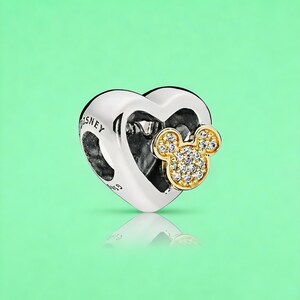May include: A silver heart-shaped charm featuring a gold-toned Mickey Mouse icon encrusted with clear stones on the front. The side of the charm is engraved with the text DISNEY and S925, showcasing a polished metallic finish and a hollow center.