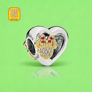 May include: A silver heart-shaped charm bead featuring a gold-toned Minnie Mouse silhouette at the center. The character design includes red stone accents on the bow and clear pav&eacute; stones on the face, set against a bright green background.