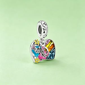 May include: A silver charm with a heart-shaped pendant featuring a colorful cartoon design. The charm depicts two characters, one blue and one pink, with a rainbow and musical notes. The charm is suspended from a loop with musical notes.