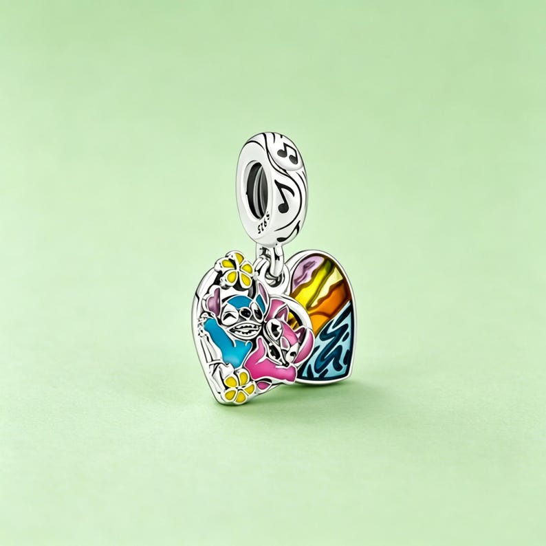 May include: A silver charm with a heart-shaped pendant featuring a colorful cartoon design. The charm depicts two characters, one blue and one pink, with a rainbow and musical notes. The charm is suspended from a loop with musical notes.