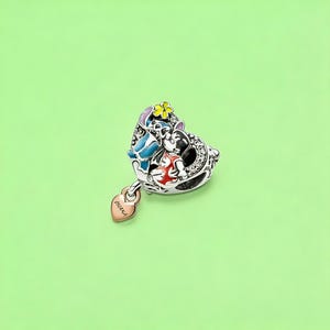 K&ouml;nnte beinhalten: Ein herzf&ouml;rmiger silberner Charm mit farbigen Emaille-Figuren von Stitch und Lilo, verziert mit einer kleinen gelben Blume und klaren Steinen. Ein baumelnder herzf&ouml;rmiger Anh&auml;nger in Ros&eacute;gold ist mit dem Wort OHANA graviert.