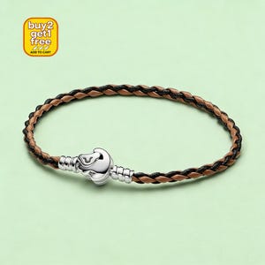 Puede incluir: Pulsera trenzada de cuero marrón y negro, con cierre plateado. La pulsera tiene forma circular y presenta un diseño tejido. El cierre es un elemento decorativo plateado. También hay un gráfico amarillo y blanco que dice "buy 2 get 1 free".