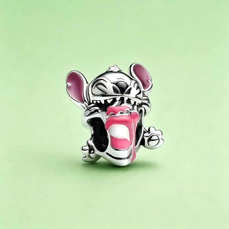 May include: A silver charm featuring the character Stitch from Lilo & Stitch. The charm depicts Stitch with pink ears and a pink and white drink. The charm is set against a light green background.