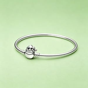 May include: A silver-colored bangle bracelet with a charm featuring a cartoon character. The charm is a stylized figure with a round body, small hat, and detailed facial features. The bracelet is on a light green background.