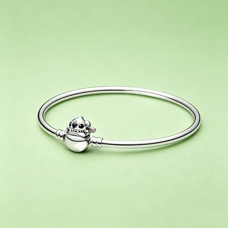 May include: A silver-colored bangle bracelet with a charm featuring a cartoon character. The charm is a stylized figure with a round body, small hat, and detailed facial features. The bracelet is on a light green background.