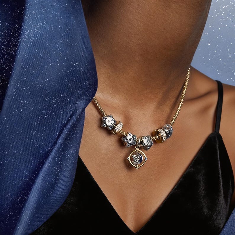 May include: A gold-toned chain necklace featuring multiple charms, including several silver-toned star-shaped beads and a central circular pendant with a rotating moon and star design, accented with small clear stones along the chain links.