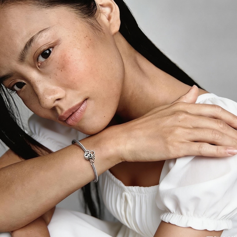 May include: A silver snake chain bracelet featuring a heart-shaped charm with a cut-out design. The charm is crafted from a combination of silver and rose gold-toned metal, secured on a flexible, textured metallic band worn around the wrist.