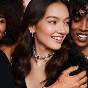 May include: A silver charm necklace featuring a variety of metallic and blue glass beads, worn around the neck. The necklace includes multiple dangling charms of different shapes and sizes, paired with small silver hoop earrings with charms.