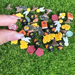 May include: A collection of miniature charms and embellishments in various shapes and colors, including hats, glasses, books, and seals. The charms are scattered on a bed of green grass, with a hand holding some of the charms.