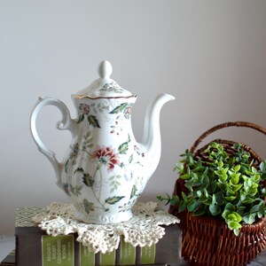 May include: White porcelain teapot with a floral pattern in red, pink, blue, and green. The teapot sits on a stack of books with a lace doily. A small wicker basket with greenery is to the right.