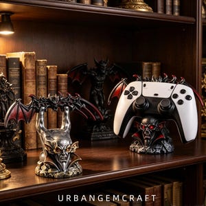 May include: A gothic-style controller stand featuring a sculpted vampire head with glowing red eyes and bat wings, designed to hold a white and black gaming controller. The base is textured like stone, and the text URBANGEMCRAFT is visible below.