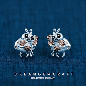 Può includere: Un paio di orecchini a forma di ape argentati. Ogni orecchino presenta un design dettagliato di ape con occhi neri, antenne e un grappolo di fiori marroni. Il testo "URBANGEMCRAFT Handcrafted Jewellery" è in basso.
