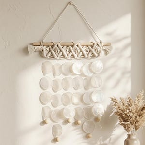 May include: A wall hanging featuring a wooden dowel with cream-coloured macrame cord knotted in a diamond pattern. Below the macrame, multiple rows of translucent, circular capiz shells are suspended, accented with small wooden beads at the ends.