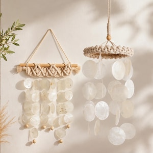 May include: A wall-hanging macrame piece with a wooden dowel and cream-coloured cord, featuring rows of circular white capiz shells. Beside it, a circular macrame mobile hangs from a cord, also adorned with multiple strands of round white capiz shells.