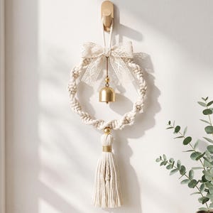 May include: A circular macrame wall hanging made of off-white twisted cotton cord. The piece features a decorative cream-colored lace bow at the top, a central gold-toned metal bell, and a long, thick tassel hanging from the bottom center.