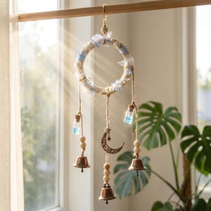 Può includere: Un ornamento circolare sospeso con un anello avvolto da accenti floreali azzurri e bianchi. Tre corde di iuta pendono dall'anello, decorate con perline di legno, piccole fiale di vetro riempite di liquido blu, campanelli in metallo e una luna.