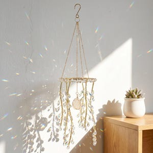May include: A hanging mobile featuring a circular gold-toned frame with multiple dangling gold wire branches adorned with clear, teardrop-shaped crystals. A single, larger, irregular-shaped pale pink crystal hangs from the center of the frame.