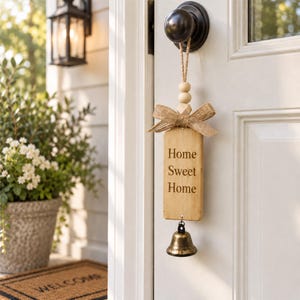 以下が含まれることがあります： ジュートロープでドアノブに吊るされた長方形の木製サイン。Home Sweet Homeの刻印入り。2つの丸い木製ビーズ、多面カットの木製ビーズ、麻の布製リボンで装飾され、下部には真鍮製のベルが取り付けられている。