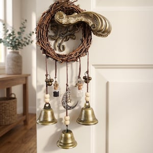 May include: A decorative door hanging featuring a circular twig wreath base with an ornate brass door handle. Suspended from the wreath are brown cords holding three brass bells, two small glass vials, wooden beads, and a crescent moon charm.