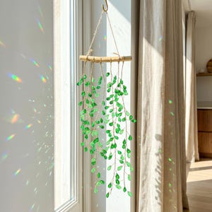Puede incluir: Una decoración de pared colgante que presenta una rama de madera horizontal de la que cuelgan varios hilos de alambre fino. Cada hilo está adornado con numerosas cuentas de cristal verde facetadas, creando un efecto en cascada.