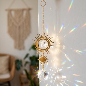 May include: A gold-toned metal suncatcher hanging from a chain, featuring a circular sunburst frame with a clear crystal star at the center, a smaller sunburst charm below, and a faceted clear crystal sphere at the bottom casting rainbow light.