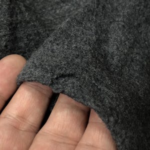 Puede incluir: Primer plano de tela gris oscuro, posiblemente de punto o jersey. La textura parece suave y ligeramente desgastada, con un borde visible donde la tela est&aacute; doblada. La imagen destaca el color y el tejido del material.