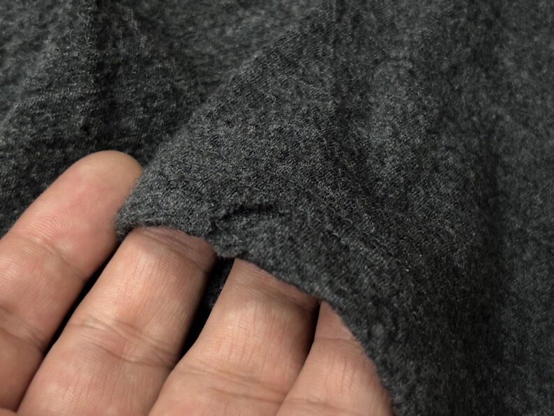Puede incluir: Primer plano de tela gris oscuro, posiblemente de punto o jersey. La textura parece suave y ligeramente desgastada, con un borde visible donde la tela est&aacute; doblada. La imagen destaca el color y el tejido del material.