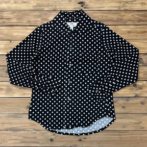 Pode incluir: Camisa preta com bolinhas brancas. A camisa de manga comprida tem gola clássica e punhos abotoados. A camisa está estendida sobre uma superfície de madeira.