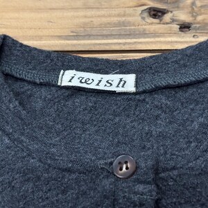 Puede incluir: Primer plano de una camiseta gris oscuro con cuello redondo y cierre de un solo bot&oacute;n. La camiseta tiene una peque&ntilde;a etiqueta blanca que dice "i wish". La tela parece ser un material de punto suave.