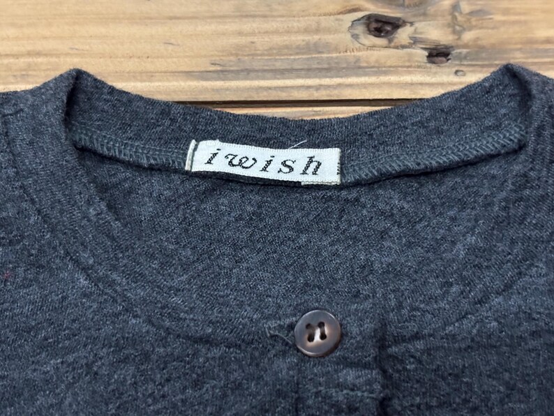 Puede incluir: Primer plano de una camiseta gris oscuro con cuello redondo y cierre de un solo bot&oacute;n. La camiseta tiene una peque&ntilde;a etiqueta blanca que dice "i wish". La tela parece ser un material de punto suave.