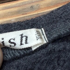 Puede incluir: Primer plano de una camiseta gris con una etiqueta blanca. La etiqueta tiene la palabra "ish" y otro texto impreso. La etiqueta tambi&eacute;n tiene las letras "YKK" impresas.