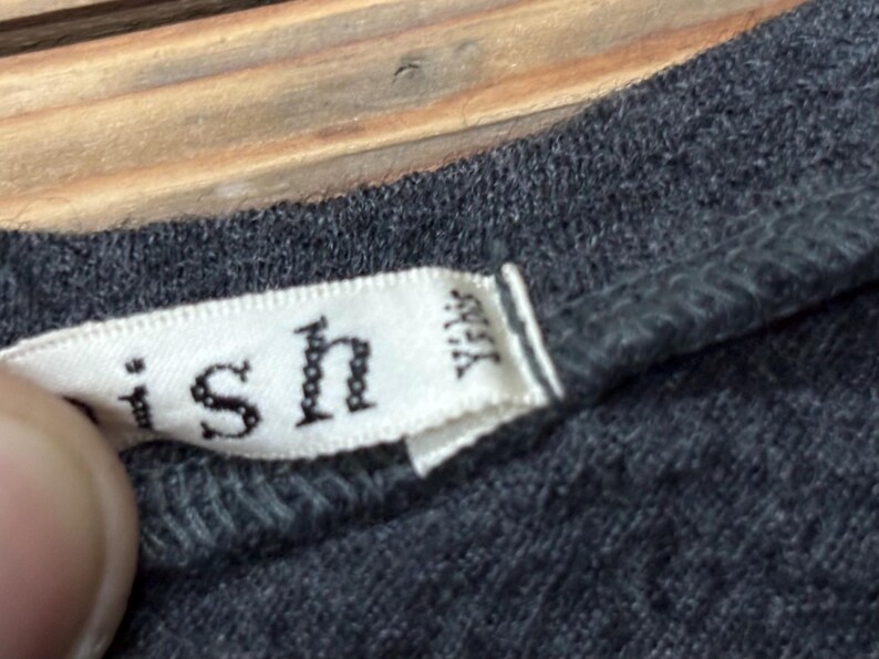 Puede incluir: Primer plano de una camiseta gris con una etiqueta blanca. La etiqueta tiene la palabra "ish" y otro texto impreso. La etiqueta tambi&eacute;n tiene las letras "YKK" impresas.