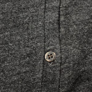 Puede incluir: Primer plano de una tela gris oscuro con un solo bot&oacute;n redondo marr&oacute;n. El bot&oacute;n est&aacute; centrado en la imagen, con la textura de la tela y las l&iacute;neas de costura verticales visibles. La tela parece ser un material de punto o jersey.