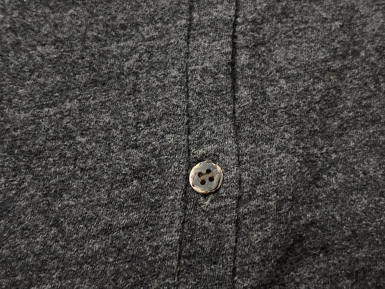 Puede incluir: Primer plano de una tela gris oscuro con un solo bot&oacute;n redondo marr&oacute;n. El bot&oacute;n est&aacute; centrado en la imagen, con la textura de la tela y las l&iacute;neas de costura verticales visibles. La tela parece ser un material de punto o jersey.