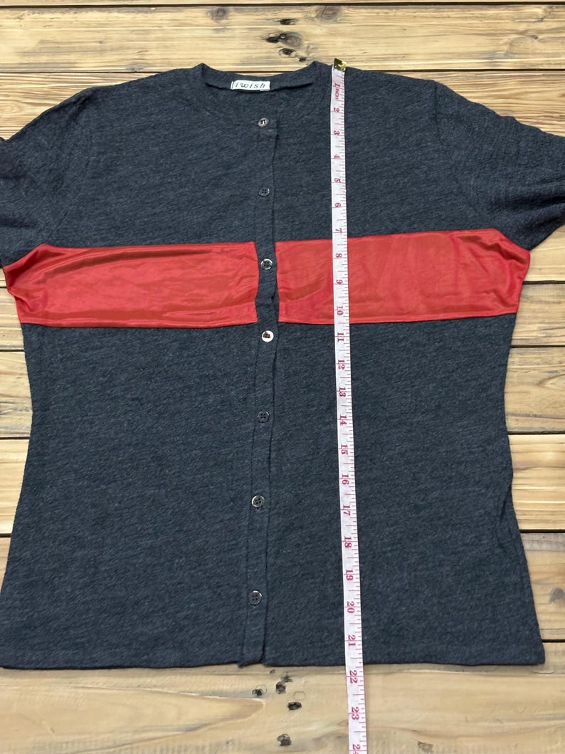 Puede incluir: Una camisa gris oscuro con botones y una franja roja horizontal en el pecho. La camisa tiene mangas cortas y una cinta m&eacute;trica se coloca verticalmente en la parte delantera. La camisa tiene la palabra "wish" impresa en la parte superior.