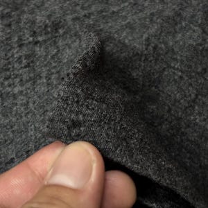 Puede incluir: Primer plano de una tela gris oscuro, posiblemente de punto o jersey. La textura parece suave y ligeramente moteada, con un brillo sutil. La tela se levanta, revelando su grosor y ca&iacute;da.