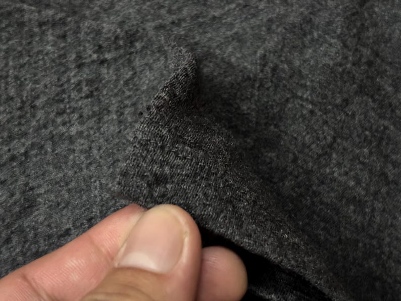 Puede incluir: Primer plano de una tela gris oscuro, posiblemente de punto o jersey. La textura parece suave y ligeramente moteada, con un brillo sutil. La tela se levanta, revelando su grosor y ca&iacute;da.