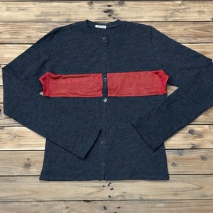 Puede incluir: Un c&aacute;rdigan gris oscuro con una franja roja horizontal en el pecho. La camisa abotonada de manga larga est&aacute; tendida sobre una superficie de madera. El c&aacute;rdigan tiene un cuello redondo y una peque&ntilde;a etiqueta cerca del cuello.