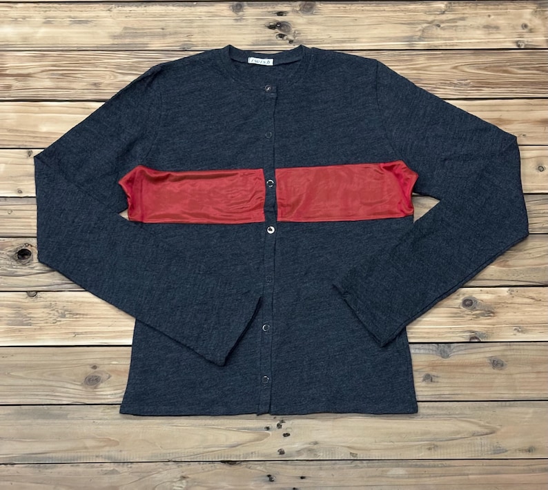 Puede incluir: Un c&aacute;rdigan gris oscuro con una franja roja horizontal en el pecho. La camisa abotonada de manga larga est&aacute; tendida sobre una superficie de madera. El c&aacute;rdigan tiene un cuello redondo y una peque&ntilde;a etiqueta cerca del cuello.