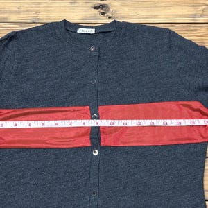 Puede incluir: Camisa abotonada gris oscuro con una franja roja horizontal en el pecho. La camisa se mide con una cinta m&eacute;trica, mostrando una medida de aproximadamente 40 cm en la franja roja.