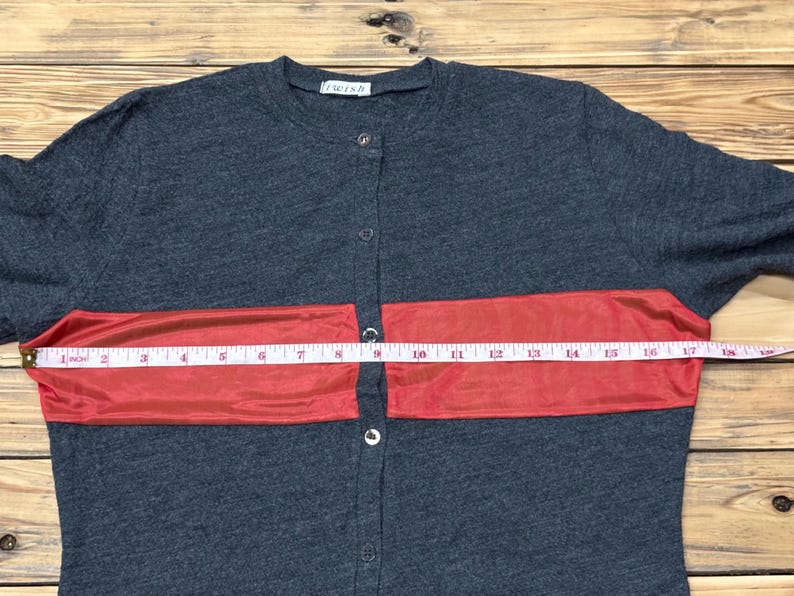 Puede incluir: Camisa abotonada gris oscuro con una franja roja horizontal en el pecho. La camisa se mide con una cinta m&eacute;trica, mostrando una medida de aproximadamente 40 cm en la franja roja.